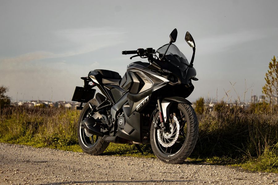 Une moto de sport noire est stationnée sur un chemin de terre en bordure de route, avec une ville en arrière-plan sous un ciel gris.